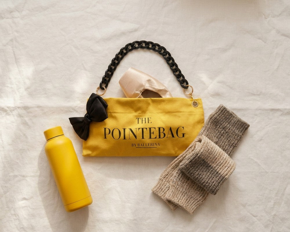The PointeBag by Ballerina - yellow cotton pointe shoe bag with black bow and chain handle, styled with ballet pointe shoes, water bottle and leg warmers